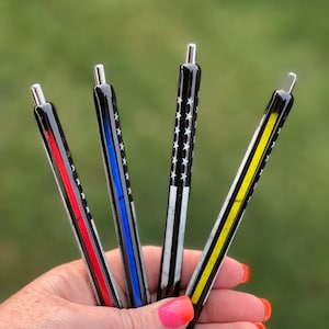 May include: Four black pens with different colored stripes and a white and black American flag design. The stripes are red, blue, yellow, and black.