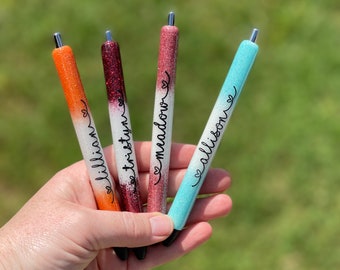 May include: Four glitter pens with personalised names: "Lillian", "Tristyn", "Macalor", and "Allison". The pens are ombre with white at the top and a gradient of colour to the bottom. The pens are orange, red, pink, and blue.