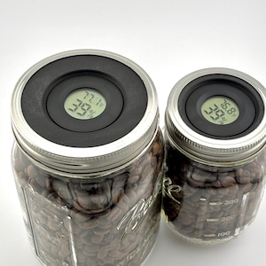 May include: Two glass jars filled with coffee beans, both with black lids that have digital humidity and temperature gauges. The larger jar reads 77.1 degrees Fahrenheit and 55% humidity. The smaller jar reads 76.8 degrees Fahrenheit and 50% humidity.