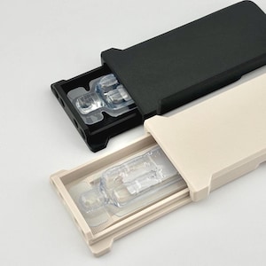 May include: Two rectangular cases, one black and one beige, each with a sliding tray. The trays are partially extended, revealing clear plastic packaging containing small, clear objects. The cases are on a white surface.