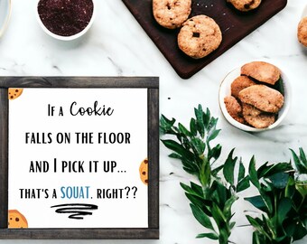 May include: Overhead shot of chocolate chip cookies on a wooden board and in bowls, with a sign that reads "If a Cookie falls on the floor and I pick it up... that's a squat, right??" The sign is framed in dark wood.