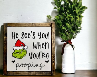 May include: A wooden sign with a white background and a brown frame. The sign features a cartoon image of the Grinch wearing a Santa hat and the text "He sees you when you're pooping".