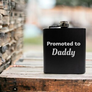 May include: Black metal flask with a silver cap and the text "Promoted to Daddy" engraved on the front.