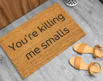 May include: A rectangular doormat made of natural fibers with the phrase "You're killing me smalls" in black lettering. Two pairs of tan sandals with ankle straps are next to the mat. The mat is on a gray wooden floor.