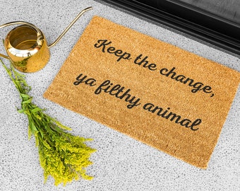 May include: A rectangular doormat made of natural fibers with the phrase "Keep the change, ya filthy animal" in black script. A gold watering can and a bundle of yellow flowers are next to the mat.