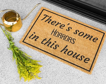 May include: A brown coir doormat with black text that reads "There's some HORRORS in this house".