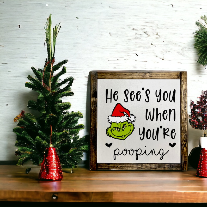 May include: A wooden sign with a white background and black text that reads "He see's you when you're pooping". The sign features a green Grinch character wearing a red Santa hat.