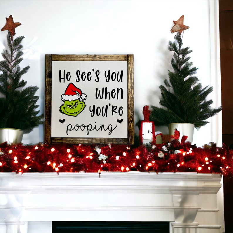 May include: A framed canvas sign with a green Grinch character wearing a Santa hat. The sign reads "He sees you when you're pooping" with hearts on either side of the word "pooping". The sign is displayed on a white fireplace mantel with a red garland and two small Christmas trees.