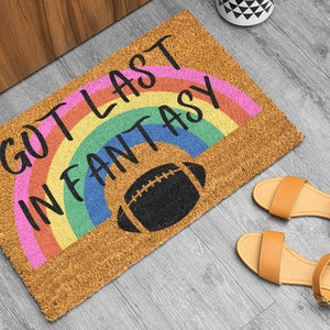 May include: A brown coir doormat with a rainbow design and the text "I got last in fantasy" in black. A black football graphic is in the center of the doormat.
