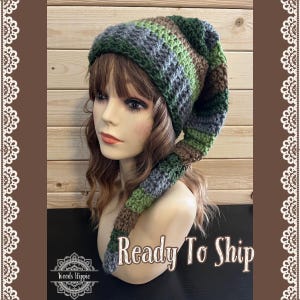 May include: A handmade crochet hat with a long, pointed end. The hat features stripes of green, gray, and brown. The text "Ready To Ship" is displayed below the hat. The hat is displayed on a mannequin head.