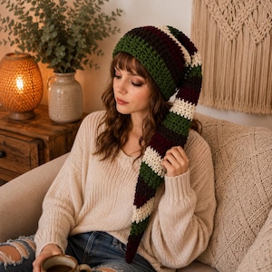 May include: A long, slouchy crochet beanie featuring horizontal stripes in dark green, deep burgundy, and cream. The hat has a tapered, extended design that hangs down to the side, crafted with a textured yarn stitch pattern throughout.