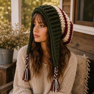 May include: A chunky crochet hat featuring a dark green ribbed border and a body with horizontal stripes in cream, dark red, and dark green. The hat includes two long side flaps that end in braided tassels made of cream, dark red, and dark green yarn.