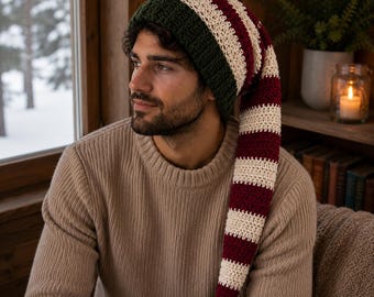 May include: A long, slouchy crochet hat featuring a dark green ribbed brim and a body with alternating horizontal stripes in deep burgundy and cream. The hat has a tapered, elongated shape that hangs down to the side, crafted from yarn.