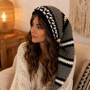 May include: A long, slouchy crochet beanie featuring a striped pattern in shades of charcoal gray, black, and white. The hat has a thick, textured knit construction with a wide brim and a long, trailing tail that hangs down the side of the head.