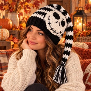 May include: A long, slouchy crochet beanie featuring a black and white striped pattern. The hat includes a large, circular white patch on the side with a black stitched skull face design and a long, dangling tail finished with a black and white tassel.