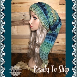 以下が含まれることがあります： 手作りのかぎ針編みの帽子、長く尖った尾が付いています。帽子は緑、青、ベージュの糸を組み合わせています。長いブロンドの髪のマネキンの頭に帽子が飾られています。「Ready To Ship」の文字が見えます。