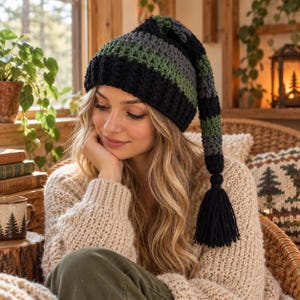 May include: A long, slouchy crochet beanie featuring horizontal stripes in black, dark gray, and olive green. The hat has a ribbed brim and a long, dangling tail that ends in a thick, black yarn tassel, creating a distinct, elongated silhouette.