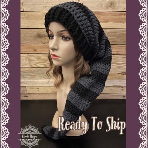 May include: A handmade crochet hat in black and gray, featuring a long, striped tail. The hat is displayed on a mannequin head. The text "Ready To Ship" is visible in the image. The hat is a unique accessory.