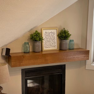 May include: A wooden shelf with a framed print that reads "There is a gathering of light, where laughter is always, a deliciousness of days, all making beautiful, the days of our lives, a gathering of sweet songs, a symphony of memories, a tapestry of laughter, a collage of happy days, a mosaic of love and peace, we love these moments". The shelf is decorated with three glass jars and two potted plants.