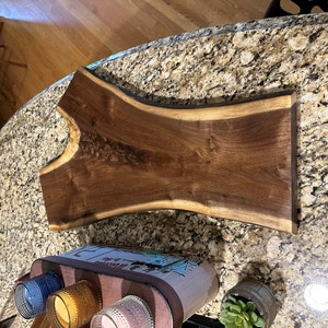 Puede incluir: Una bandeja de servir de madera marrón oscura grande con un borde natural. La bandeja está hecha de madera de nogal y tiene un acabado liso y pulido.