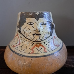 Op de afbeelding: Een afgeronde keramische pot met een brede basis en een smallere hals. Het bovenste gedeelte is voorzien van een handgeschilderd gezicht met zwart haar, amandelvormige ogen en een geometrisch patroon in zwart en roodachtig oranje.