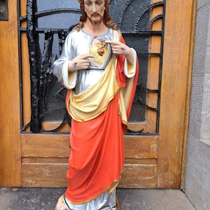 Op de afbeelding: Een standbeeld van Jezus Christus, gekleed in een witte en gouden jurk met een rode jas. Het beeld heeft een halo en een zichtbaar hart. Het beeld staat voor een houten deur en een decoratief raam.