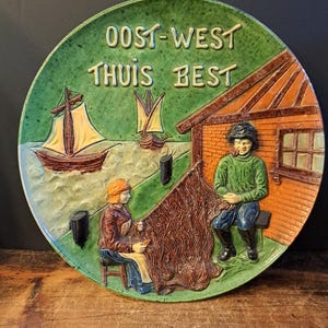 Pode incluir: Prato de cerâmica com borda verde e cena central em relevo. A cena retrata dois veleiros na água, um pescador, uma mulher e um edifício. O prato tem o texto "OOST-WEST THUIS BEST".