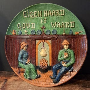 Pode incluir: Prato de cerâmica com borda verde e o texto "EIGEN HAARD IS GOUD WAARD". O prato retrata um casal sentado perto de uma lareira, com uma chaleira pendurada sobre as chamas. O prato é marrom, verde e azul.