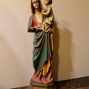 Op de afbeelding: Handgeschilderd houten beeld van de Maagd Maria met een kind. Het beeld heeft een kleurenpalet van rood, groen, geel en bruin. De basis is een donkerbruine, achthoekige vorm.