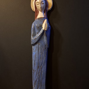 Puede incluir: Una figurita de cerámica de una mujer con una túnica azul con las manos juntas en oración. Tiene un halo y está de pie sobre una superficie de madera.