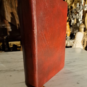 Peut inclure: Un livre relié en cuir rouge foncé avec un marque-page en ruban rouge. Le livre est posé sur une surface en marbre blanc.