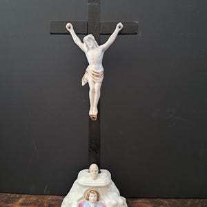 May include: A religious figurine featuring a white Jesus Christ figure on a black wooden cross. The base is a white porcelain structure with an angel and skull design. The figurine is set against a dark background.