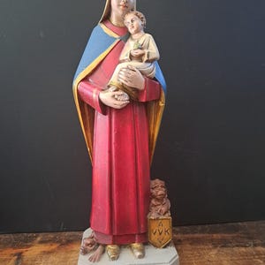 Op de afbeelding: Een vintage beeld van een vrouw die een kind vasthoudt, gekleed in rode en blauwe gewaden. Het beeld heeft een lichtgekleurd gezicht en staat op een grijze basis. Een kleine leeuw en schild staan aan de basis.