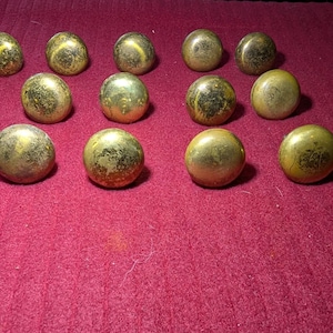 May include: A collection of antique brass knobs with a weathered appearance. The knobs are round with a domed top and varying shades of gold and green. They are arranged on a deep red surface.