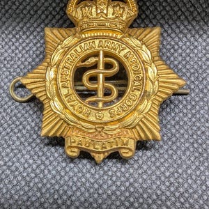 May include: A gold-coloured Royal Australian Army Medical Corps badge. The badge features a central caduceus symbol, surrounded by a laurel wreath and a crown at the top. The text "ROYAL AUSTRALIAN ARMY MEDICAL CORPS" is inscribed.