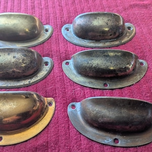 May include: Six antique brass drawer pulls with a weathered finish. Each pull has a curved handle and a backplate with two screw holes. The pulls are in various shades of gold and bronze, showing signs of age and use.
