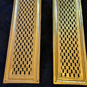 May include: Two antique brass rectangular vent covers. Each cover features a diamond-shaped lattice pattern within a rectangular frame. The top of each cover has a decorative arched design. The brass has a warm, aged patina.