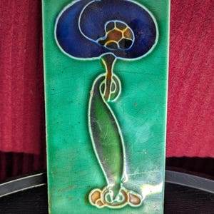 May include: Rectangular ceramic tile with a floral design. The tile features a blue flower with a brown center and a green stem and leaves against a green background. The tile is approximately 10 cm wide and 20 cm tall.