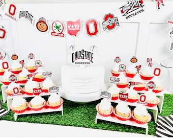 May include: A festive display featuring a white cake and cupcakes, all decorated with Ohio State Buckeyes themed toppers and banners. The cupcakes are arranged on tiered white stands, with red and white accents.