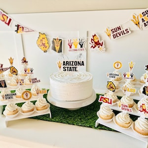May include: A white frosted cake and cupcakes decorated with Arizona State University (ASU) themed toppers and banners. The cake has a "ARIZONA STATE" cake topper. The cupcakes are arranged on tiered stands, with ASU logos and "SUN DEVILS" text.
