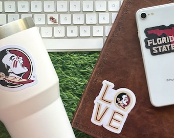May include: A white tumbler with a Florida State Seminoles logo sticker. The tumbler is sitting on a green surface next to a keyboard and a brown leather notebook. The notebook has a Florida State Seminoles sticker and a "LOVE" sticker with a Seminole logo.