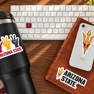 May include: A black insulated tumbler featuring a sticker with three hand signs in maroon, gold, and black with the text Arizona State. A silver smartphone displays a trident sticker, and a second sticker with a trident and Arizona State text.