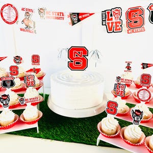 May include: A white frosted cake with a red and black "NC State" cake topper, surrounded by cupcakes with matching toppers. The scene is decorated with NC State Wolfpack banners and stickers, creating a festive atmosphere.