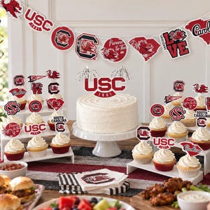 May include: A party decoration set featuring University of South Carolina themed items, including a banner with USC and Gamecocks logos, a large cake topper, and numerous cupcake toppers with various team graphics, all in garnet and white colors.