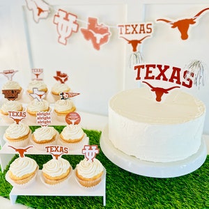 May include: A white cake decorated with a brown and white Texas Longhorns logo topper. The cake is surrounded by cupcakes decorated with various Texas Longhorns logos and sayings, including "Hook 'em Horns", "Texas Longhorns", and "Alright Alright Alright".