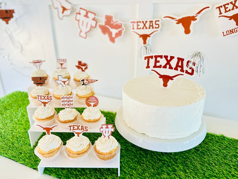 Peut inclure: Un g&acirc;teau blanc d&eacute;cor&eacute; d'un topper repr&eacute;sentant le logo des Texas Longhorns en brun et blanc. Le g&acirc;teau est entour&eacute; de cupcakes d&eacute;cor&eacute;s de diff&eacute;rents logos et slogans des Texas Longhorns, notamment "Hook 'em Horns", "Texas Longhorns" et "Alright Alright Alright".