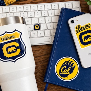 May include: A white stainless steel tumbler, a silver smartphone, and a blue notebook, each featuring University of California, Berkeley themed stickers. The tumbler has a shield-shaped sticker with a bear and the text California. The phone and notebook display bear and Cal logo stickers.