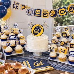 May include: A party table decorated with University of California, Berkeley themed items including a white frosted cake with a Cal bear paw logo topper, cupcakes with various Cal logo picks, blue and yellow pennant banners, and Cal napkins.