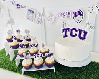 May include: A white cake with purple frosting and a "TCU" topper, surrounded by cupcakes with purple frosting and various TCU themed toppers. The cupcakes are on a white tiered stand. The background is a green grass surface.