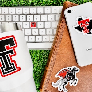 May include: A white tumbler, a white iPhone, and a keyboard are arranged on a bed of green grass. The tumbler and phone feature a red and black logo. A sticker of a rider on a horse is also visible.